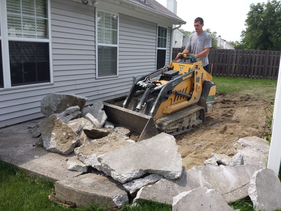 A small skid loader is demolishing and removing a previous patio to prepare for a replacement one. 