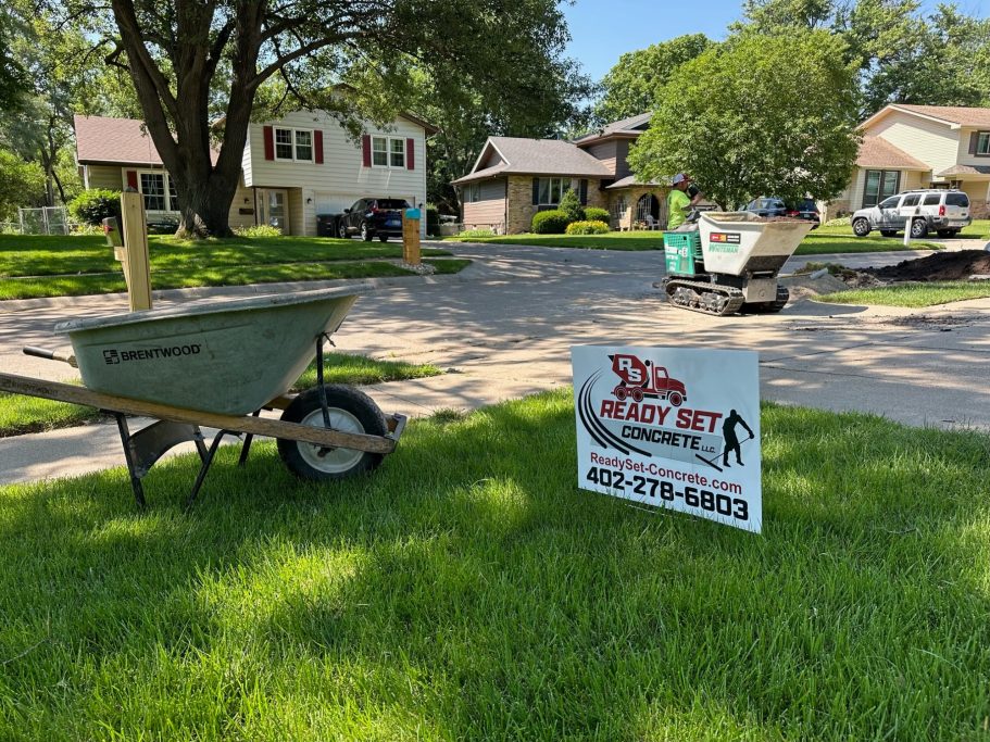 Yard with a sign reading 'Ready Set Concrete' and wheelbarrow filled with concrete material in the background 