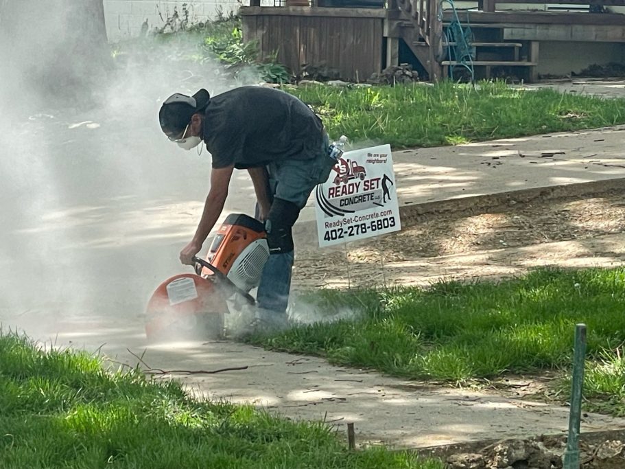 Worker drilling into concrete to prepare for a new sidewalk, surrounded by dust and debris. The worker is using a power tool